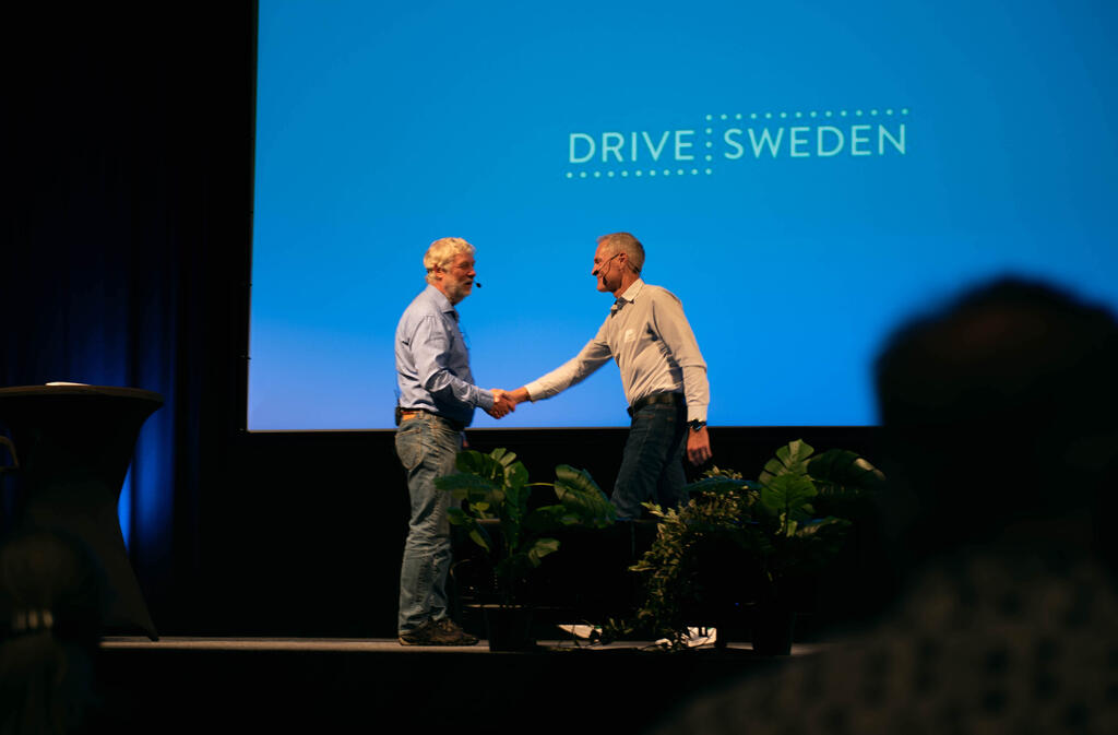 Två män skakar han på en scen. Drive Swedens logotyp på skärmen i bakgrunden